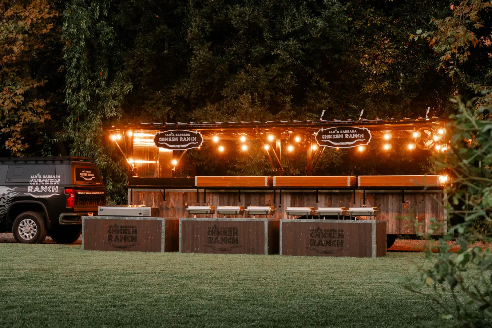 The Santa Barbara Chicken Ranch grill trailer set up on a lawn at dusk, strung with lights and lined with buffet stations