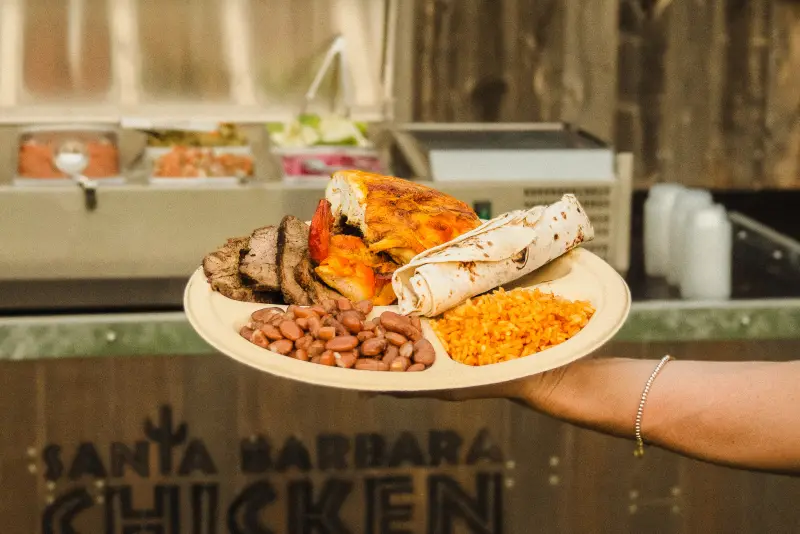 A plate of tri-tip, grilled chicken, beans, rice and a flour tortilla held up in front of a Chicken Ranch buffet station