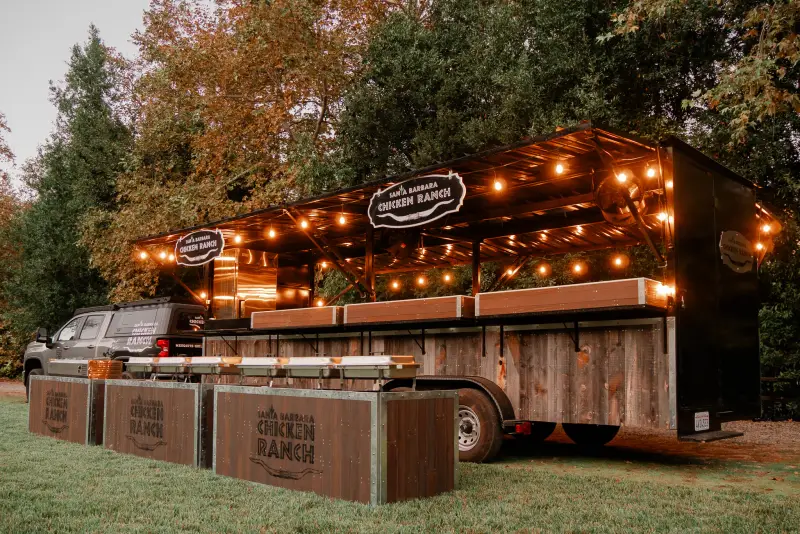 The grill trailer from the side at dusk, lit under its awning, with buffet stations lined up in front